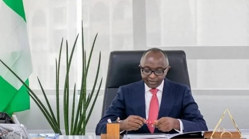 Nigerian government official seated at a desk in an office, with a Nigerian flag visible in the background.